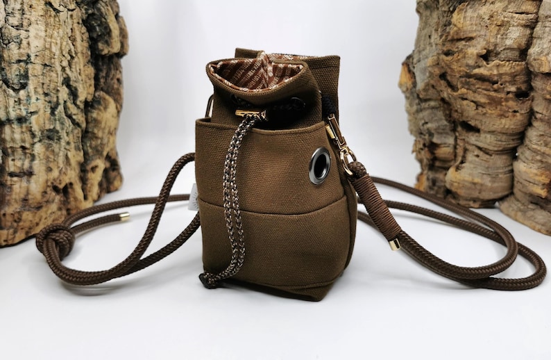 Könnte beinhalten: Eine braune Canvas-Tasche mit Kordelzugverschluss und gemustertem Innenraum. Die Tasche hat einen geflochtenen Kordelzug und einen langen, verstellbaren Schulterriemen. Die Tasche ist mit einem kleinen, abgerundeten Boden und Metallösen versehen.