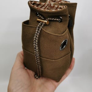 Puede incluir: Bolsa de lona marr&oacute;n con cord&oacute;n y un interior estampado en contraste. La bolsa presenta un detalle met&aacute;lico dorado y un cord&oacute;n trenzado. Tiene un peque&ntilde;o bolsillo y un ojal met&aacute;lico. Altura aproximada: 15 cm.