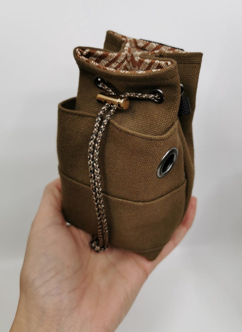 Puede incluir: Bolsa de lona marr&oacute;n con cord&oacute;n y un interior estampado en contraste. La bolsa presenta un detalle met&aacute;lico dorado y un cord&oacute;n trenzado. Tiene un peque&ntilde;o bolsillo y un ojal met&aacute;lico. Altura aproximada: 15 cm.