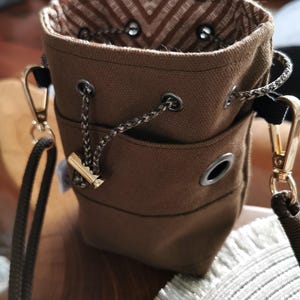 Puede incluir: Un bolso tipo cubo de lona marr&oacute;n con cierre de cord&oacute;n y herrajes dorados. El bolso tiene un forro interior estampado y una correa para el hombro desmontable. El bolso mide aproximadamente 18 cm de alto.