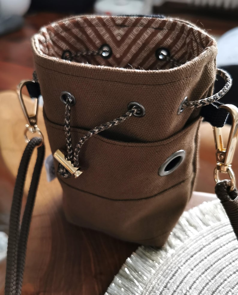 Puede incluir: Un bolso tipo cubo de lona marr&oacute;n con cierre de cord&oacute;n y herrajes dorados. El bolso tiene un forro interior estampado y una correa para el hombro desmontable. El bolso mide aproximadamente 18 cm de alto.