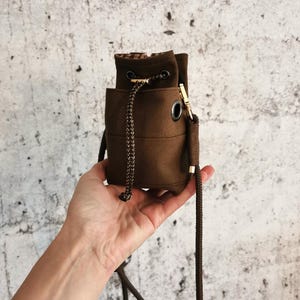 Puede incluir: Un peque&ntilde;o bolso de cubo de lona marr&oacute;n con cierre de cord&oacute;n y una correa larga y ajustable. El bolso presenta herrajes met&aacute;licos de color dorado y un forro interior estampado. El bolso est&aacute; en la mano de una persona.