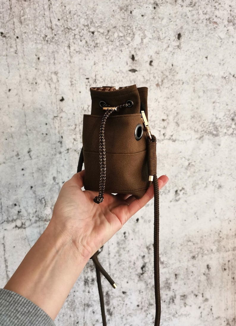 Puede incluir: Un peque&ntilde;o bolso de cubo de lona marr&oacute;n con cierre de cord&oacute;n y una correa larga y ajustable. El bolso presenta herrajes met&aacute;licos de color dorado y un forro interior estampado. El bolso est&aacute; en la mano de una persona.