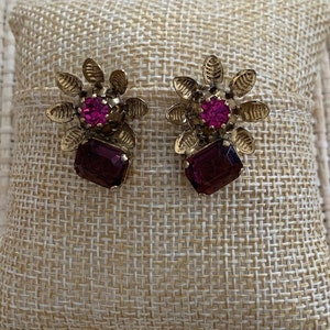 May include: A pair of gold tone earrings with a flower design and a purple gem in the center.