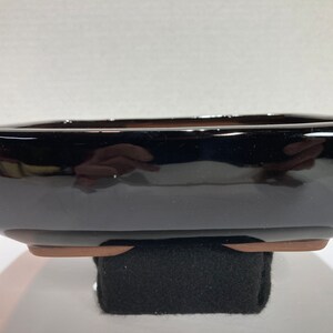 May include: A black square ceramic bonsai pot with a brown rim and a glossy finish. The pot is sitting on a black surface.