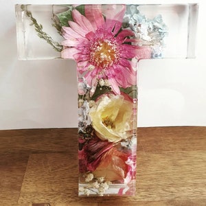 May include: A clear glass letter T with pressed flowers inside. The flowers are pink, white, and yellow. The letter is a decorative piece for a home or office.
