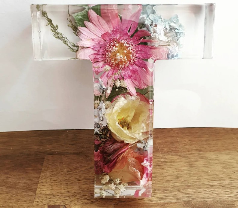 May include: A clear glass letter T with pressed flowers inside. The flowers are pink, white, and yellow. The letter is a decorative piece for a home or office.