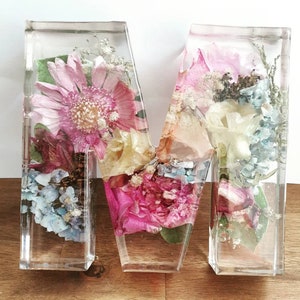 May include: A clear resin letter M filled with pressed flowers in shades of pink, white, and blue. The flowers are arranged in a random pattern and create a beautiful, delicate design.
