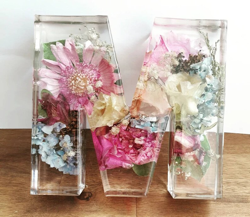 May include: A clear resin letter M filled with pressed flowers in shades of pink, white, and blue. The flowers are arranged in a random pattern and create a beautiful, delicate design.