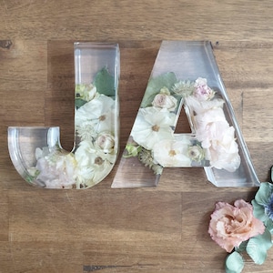 May include: Two clear acrylic letters, "JA", filled with pressed flowers in shades of white, pink, and green. The letters are sitting on a wooden surface.