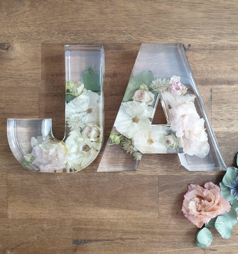 May include: Two clear acrylic letters, "JA", filled with pressed flowers in shades of white, pink, and green. The letters are sitting on a wooden surface.