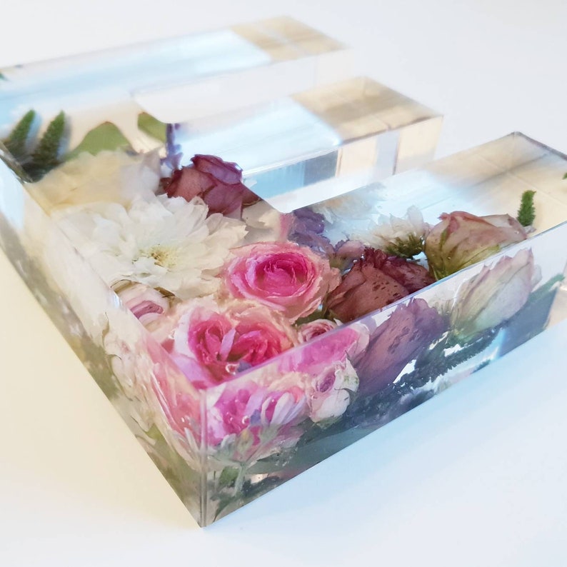 May include: A clear acrylic letter E filled with pressed flowers. The flowers are pink, white, and purple.