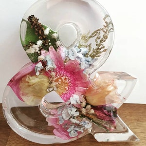 May include: A clear resin ampersand with pressed flowers and greenery inside. The flowers are pink, yellow, and white.