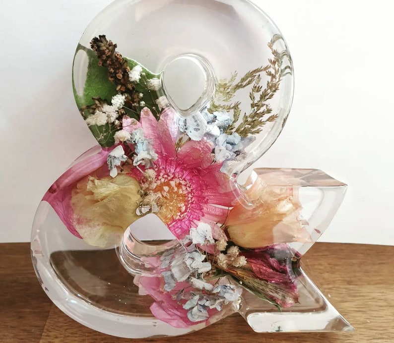 May include: A clear resin ampersand with pressed flowers and greenery inside. The flowers are pink, yellow, and white.