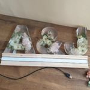 May include: A wedding sign with the letters A & J made of clear acrylic with flowers inside. The letters are lit with white lights and are sitting on a wooden base.