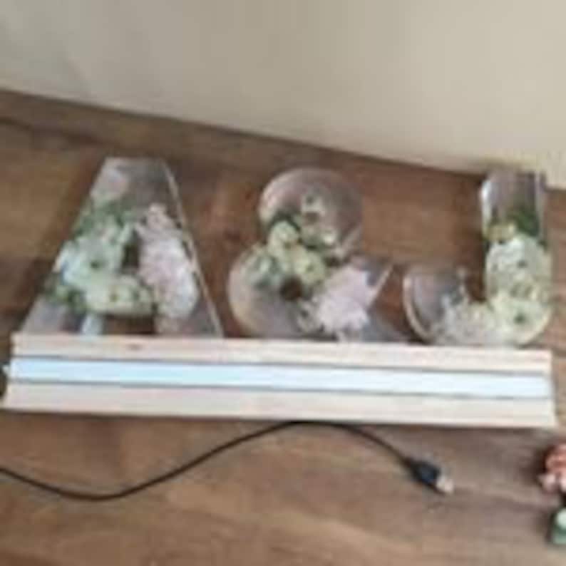 May include: A wedding sign with the letters A & J made of clear acrylic with flowers inside. The letters are lit with white lights and are sitting on a wooden base.