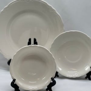 May include: Three white ceramic plates with scalloped edges, displayed on black stands. The plates are different sizes, with the largest plate in the back and the smallest plate in the front.