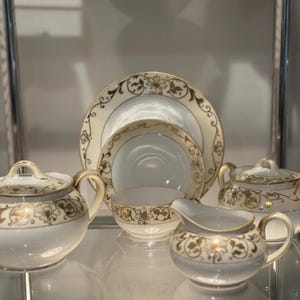 以下が含まれることがあります： 金色の花のディテールが施されたクリーム色のティーセット。ティーポット、砂糖入れ、クリーマー、いくつかのプレートとボウルが含まれています。各ピースの縁は金色でトリミングされています。