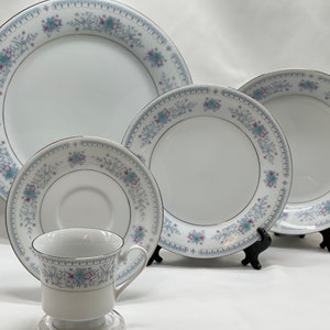 May include: A set of white porcelain dinnerware with a delicate blue and purple floral pattern. The set includes a large dinner plate, a salad plate, a saucer, and a teacup.