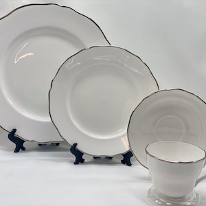 Op de afbeelding: Een set wit porselein serviesgoed met een zilveren rand. De set bevat een groot dinerbord, een kleiner saladebord, een schoteltje en een theekopje.