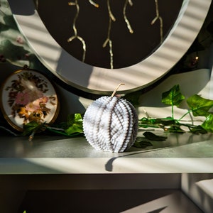 May include: A white, decorative candle with a geometric pattern sits on a white shelf in front of a mirror. The candle is surrounded by green ivy.