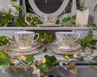 May include: Three white teacups and saucers with a gold leaf pattern. The cups and saucers are arranged on a white surface with a garland of green leaves and white lights.