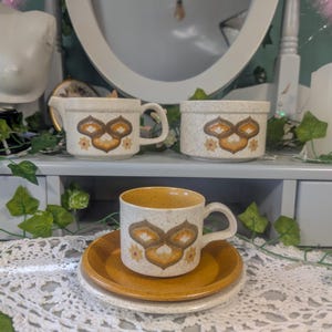Könnte beinhalten: Eine Vintage-Kaffeetasse aus Keramik mit passender Untertasse. Die Tasse und die Untertasse sind cremefarben mit einem braunen und orangefarbenen Blumendesign. Ein Sahnekännchen und eine kleine Schüssel mit dem gleichen Design sind ebenfalls sichtbar.