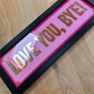 May include: A framed artwork featuring the phrase "LOVE YOU, BYE!" in gold lettering on a pink background. The frame is black. The piece is angled, suggesting it's a decorative item for a home or office, adding a touch of modern style.
