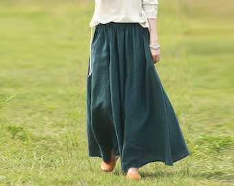 May include: A woman wearing a white long-sleeved shirt and a dark green maxi skirt. The skirt has a gathered waist and a flowing silhouette.