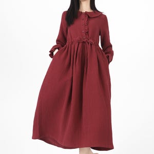 May include: A long, burgundy dress with a round collar and long sleeves. The dress features a button-down front, a cinched waist with a tie, and pockets. The dress is knee-length and has a flowing skirt. The model is wearing brown flats.