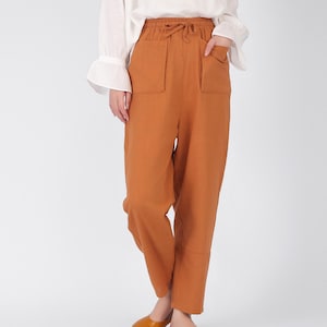 May include: Rust-coloured trousers with a drawstring waist and two large front pockets. The trousers are paired with a white long-sleeved blouse. The outfit is completed with brown leather slip-on shoes. The trousers are made of a soft, flowing fabric.