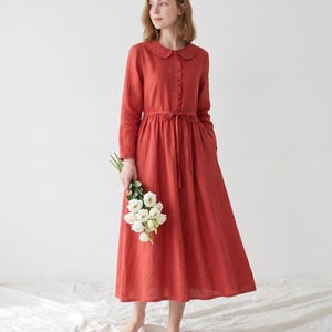 May include: A long-sleeved, ankle-length dress in a solid rust-red color. The garment features a rounded Peter Pan collar, a front button placket with a ruffled trim, and a matching fabric tie at the waist. The skirt is full and gathered.