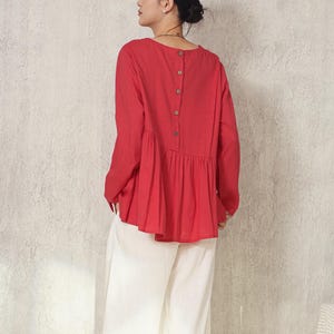 以下が含まれることがあります： 赤い長袖ブラウス、背中にボタン、裾にフリル、白いワイドパンツの組み合わせ。 シンプルでエレガントな装いで、カジュアルウェアに適しています。 背景はニュートラルな質感の壁です。