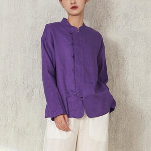 May include: A woman wearing a purple long-sleeved shirt with a mandarin collar and button closure, paired with white wide-leg trousers. The shirt is a vibrant purple, and the trousers are a light cream colour. The outfit is simple and elegant.