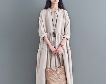 May include: A beige linen dress with a collared neckline and a gathered skirt. The dress has long sleeves and a button closure at the front. The model is wearing a brown necklace and a woven tote bag.