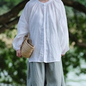 May include: A white linen blouse with a round neckline and long sleeves, paired with light green linen pants. The person is holding a small woven basket.