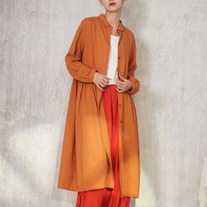May include: A long, orange-brown shirt with long sleeves, worn over a white top and paired with bright orange trousers. The outfit is completed with black shoes. The clothing has a relaxed, flowing style.