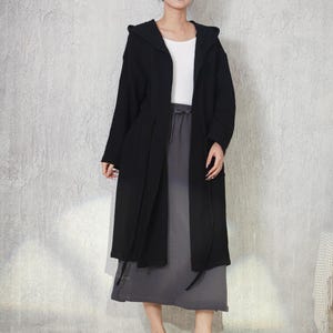Könnte beinhalten: Eine schwarze langärmelige Strickjacke mit Kapuze über einem weißen Oberteil und einem anthrazitfarbenen Midirock. Die Strickjacke hat ein offenes Design mit langen Bindebändern an der Taille, während der Rock einen Kordelzug und einen geraden Saum hat.