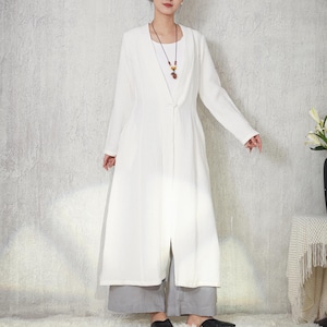 May include: A long, white, open-front coat with long sleeves and a single button closure. The coat is worn with grey wide-leg trousers and black flats. A necklace with a pendant is visible. The background is a textured, light grey wall.