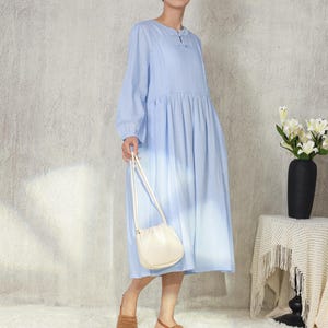 May include: A light blue long-sleeve dress with a gathered waist and a small keyhole detail at the neckline. The dress is paired with a cream-colored crossbody bag and brown woven sandals. A black vase with white flowers sits on a side table.