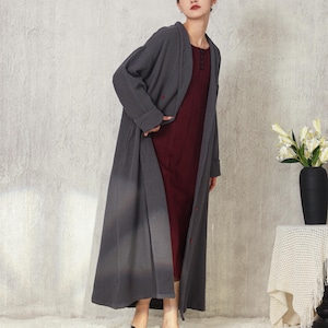 May include: A woman in a long, grey duster coat over a burgundy dress. The coat has a button closure and a subtle ombre effect. She wears black flats. A black vase with white flowers sits on a small table.