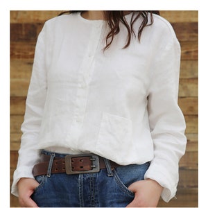 May include: A white linen shirt with a button-up front and a single pocket on the left side. The shirt is worn with a brown leather belt with a silver buckle.