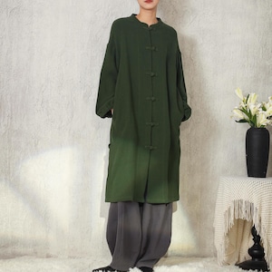 May include: An olive green coat with a mandarin collar and frog closures. The coat is worn with loose, grey trousers. The outfit is simple and elegant, suitable for casual or semi-formal occasions. The coat has long sleeves and pockets.