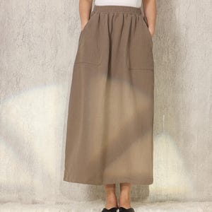 Könnte beinhalten: Eine Frau trägt ein weißes T-Shirt mit Rundhalsausschnitt und einen langen, braunen Rock mit Taschen. Der Rock hat einen dezenten Ombre-Effekt, der von einem dunkleren Farbton oben zu einem helleren Farbton unten übergeht. Schwarze Ballerinas vervollständigen das Outfit.