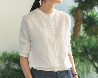 May include: A white linen shirt with a mandarin collar and long sleeves. The shirt has a button-up front and is tucked into a pair of dark blue trousers.