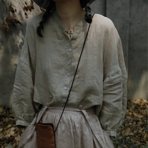 Op de afbeelding: Een beige linnen outfit bestaande uit een overhemd met knopen, een rok met zakken en een kleine bruine crossbodytas. Het overhemd heeft een opstaande kraag en lange mouwen. De rok is hoog in de taille en vastgebonden in de taille. De tas is rechthoekig.