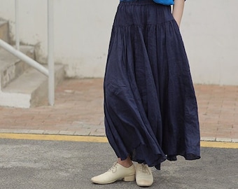 May include: A woman wearing a blue collared shirt, a wide-brimmed straw hat, a dark blue skirt, and white shoes. She is standing in front of a white wall with a white railing and steps.