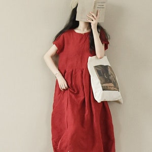 May include: A woman wearing a red linen dress with short sleeves and a round neckline. She is holding a book in front of her face and carrying a white tote bag with a printed design. She is wearing brown sandals.