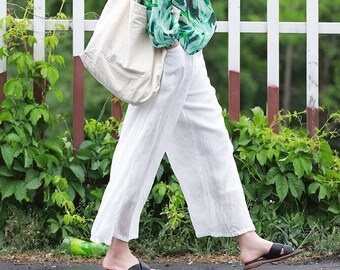 May include: A woman wearing a white linen pants and a green and white floral print blouse. She is carrying a white tote bag and walking on a sidewalk.