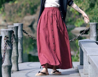 May include: A woman wearing a red skirt with a black button-down shirt and a brown cap. She is standing on a wooden bridge with a chain link fence.